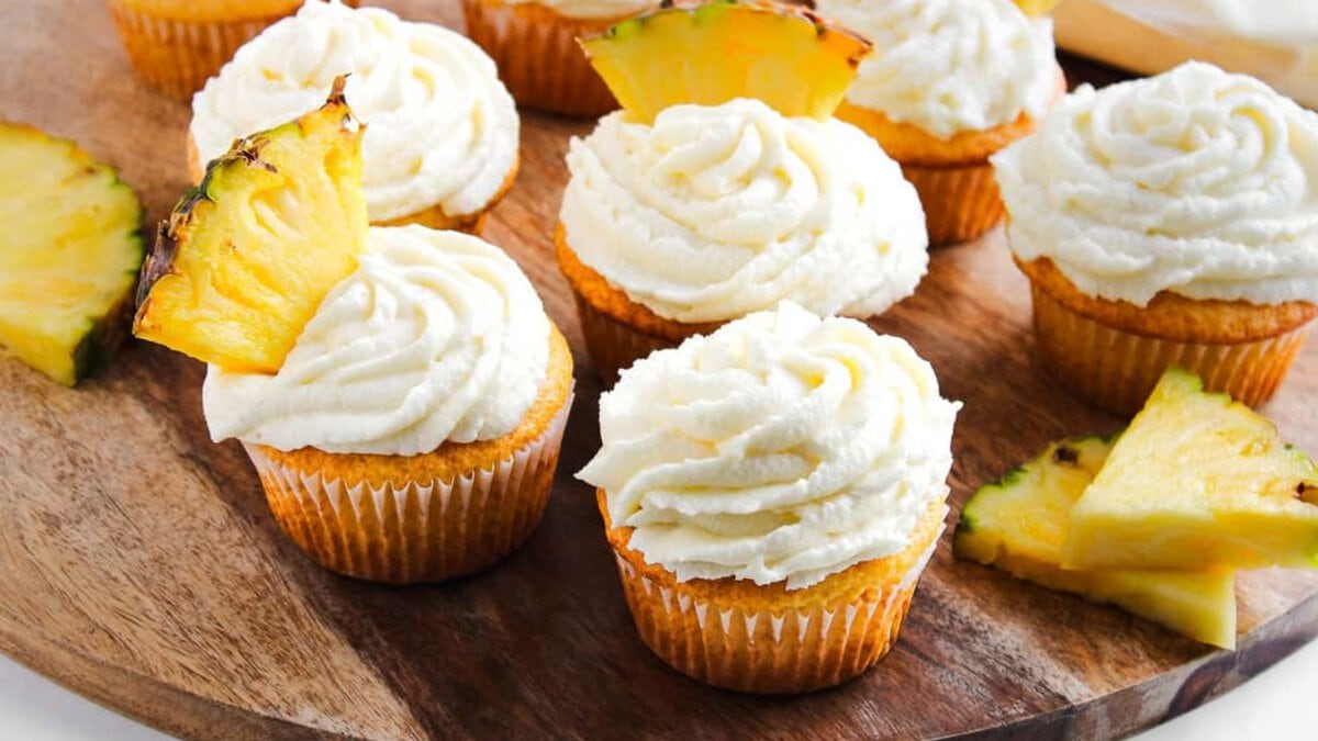 Cupcakes with swirled white frosting, some topped with pineapple slices, arranged on a wooden board.