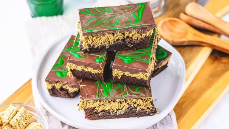 Three layered chocolate and oat bars with green swirled chocolate topping are stacked on a white plate, surrounded by kitchen utensils and ingredients.