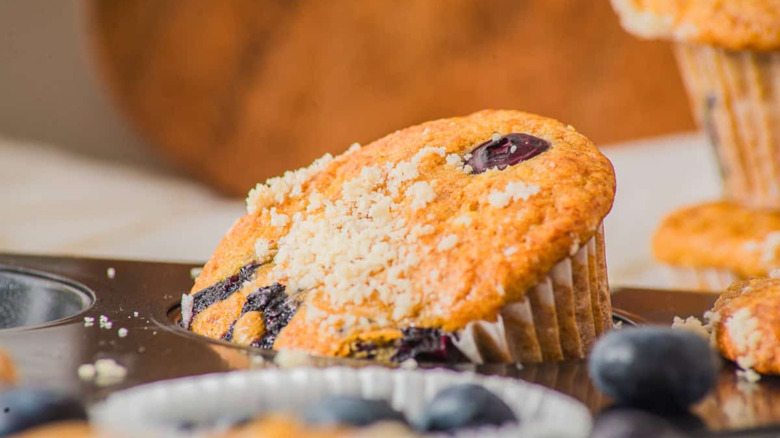 Close-up of blueberry muffins in a baking tray, with fresh blueberries and muffin crumbs scattered around.