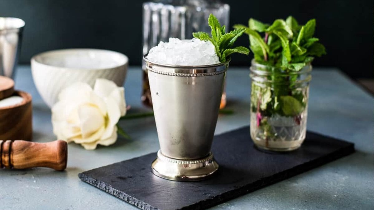 A mint julep in a silver mint julep cup with sprigs of mint and a decanter of bourbon and other ingredients in the background.
