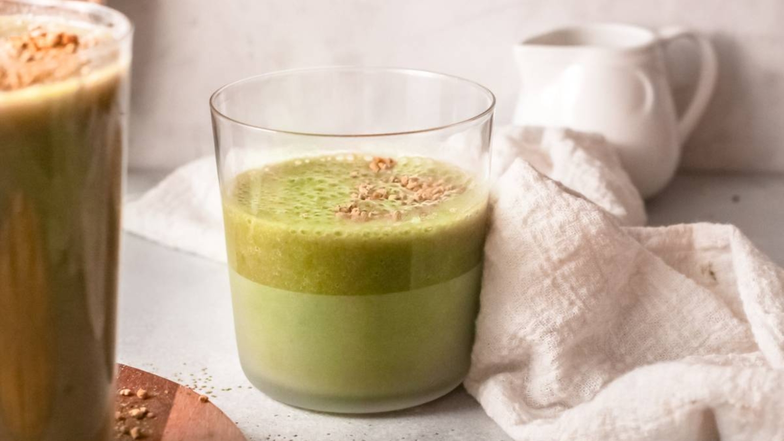 A clear glass filled with green smoothie on a white surface, next to a white cloth and a small white pitcher in the background.