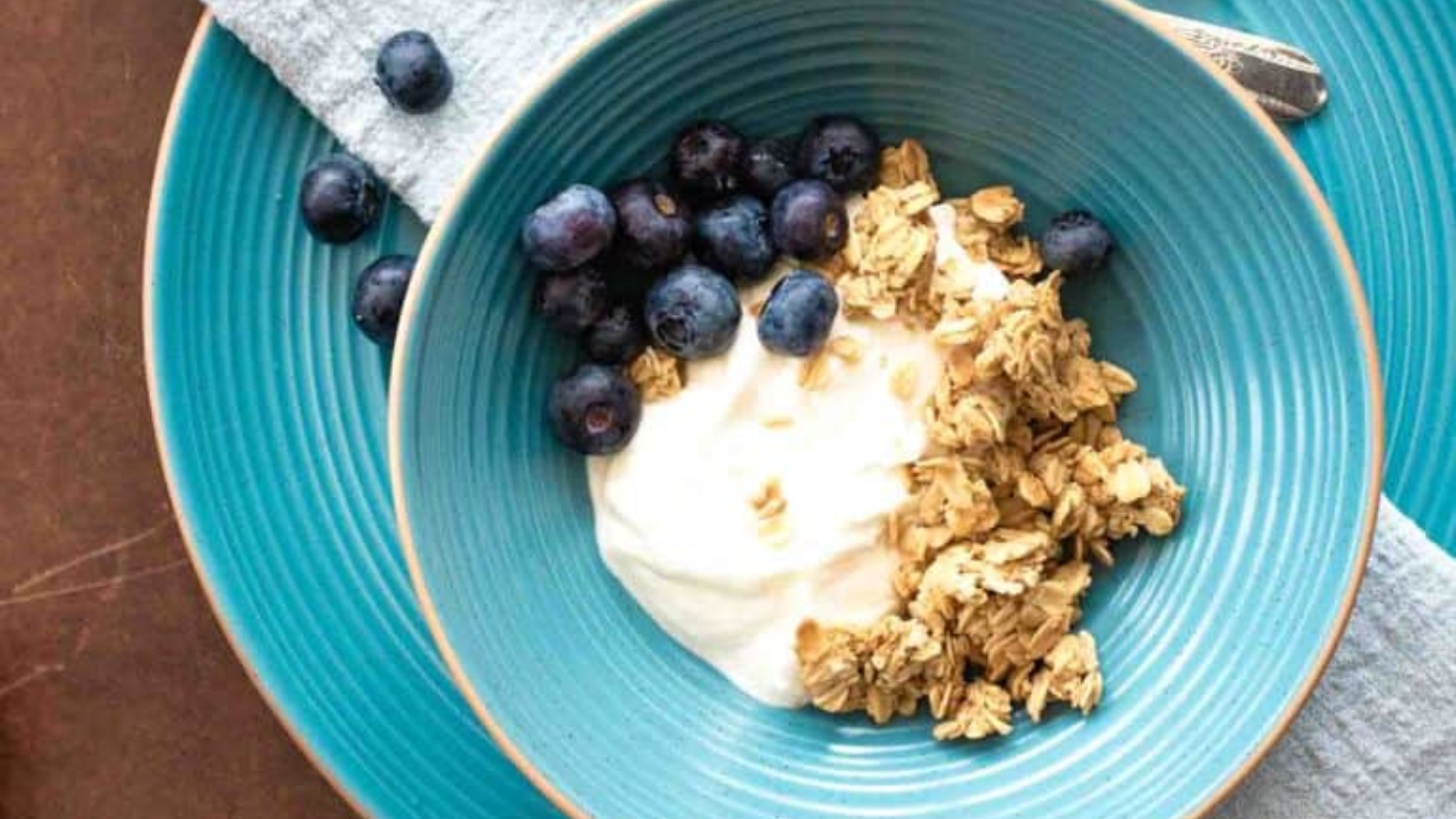 A blue bowl containing granola, blueberries, and yogurt, placed on a matching blue plate with a spoon and a folded napkin beside it&mdash;perfect for adding to your list of thoughtful Mother&rsquo;s Day recipes.