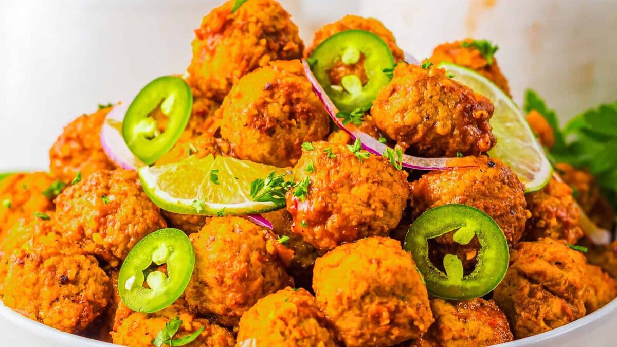 Spicy meatballs garnished with lime slices, jalape&ntilde;o rings, and red onion on a white plate.