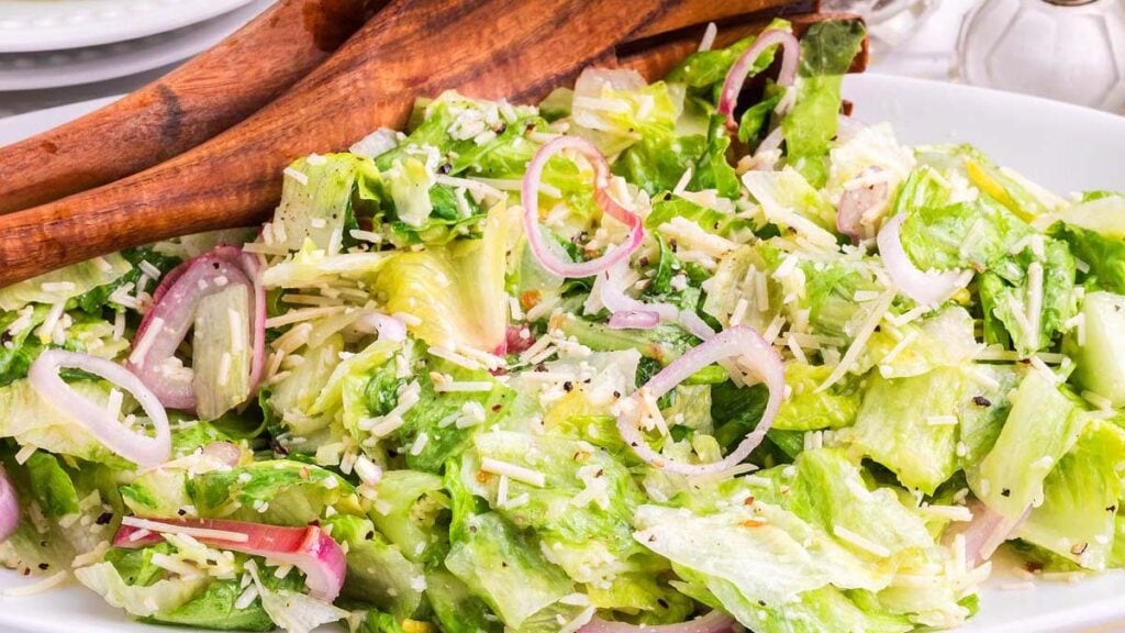 A bowl of tossed green salad with lettuce, sliced red onions, grated cheese, and wooden salad tongs.