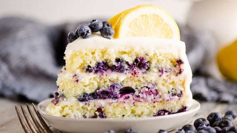 A slice of layered blueberry lemon cake with white frosting, topped with fresh blueberries and a lemon slice, on a white plate with a fork.
