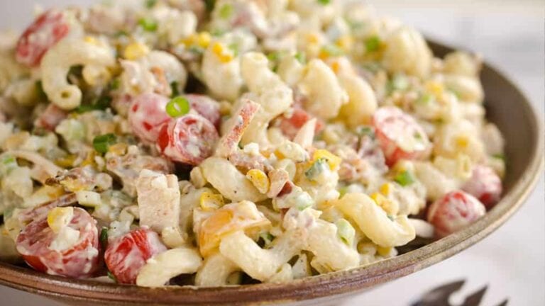 A bowl of creamy macaroni salad with cherry tomatoes, green onions, corn, and chopped vegetables.