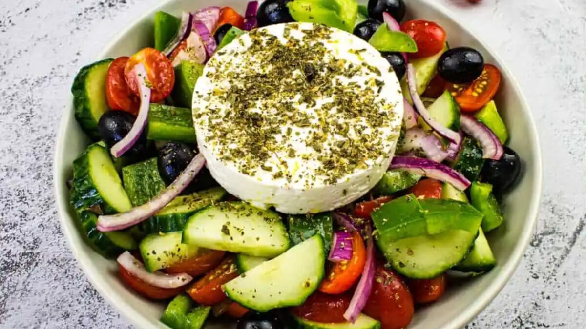 A bowl of Greek salad with cucumbers, tomatoes, red onions, black olives, and a round slice of feta cheese topped with herbs.