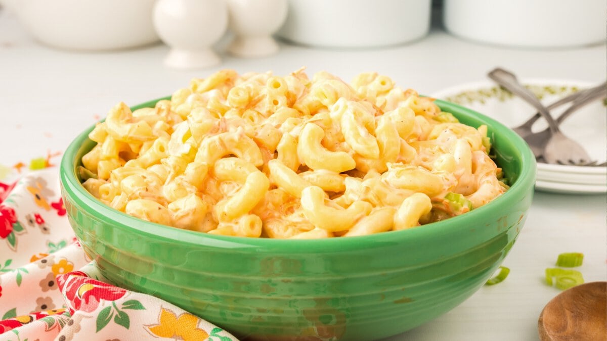 A green bowl filled with creamy macaroni and cheese sits on a table next to a floral napkin, a fork, and a spoon—an irresistible choice for Memorial Day BBQ dishes.
