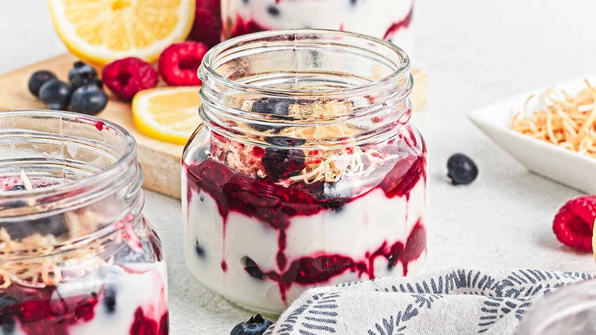 Glass jars filled with yogurt, berry sauce, and toasted coconut, with lemons and berries in the background.