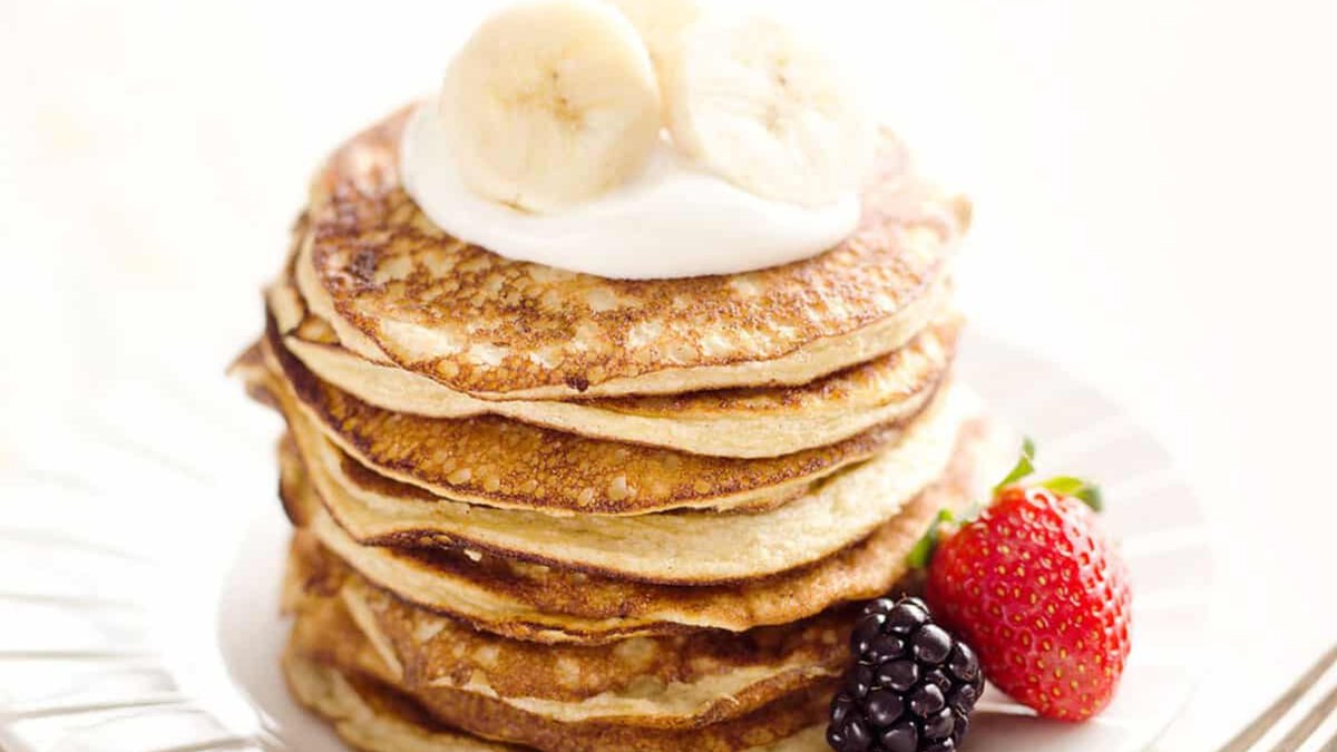 A stack of pancakes topped with banana slices and cream, served with a strawberry and a blackberry on a white plate.