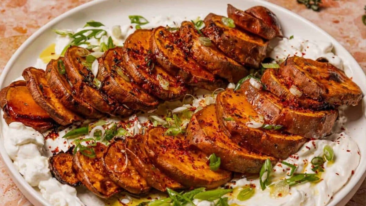 Sliced roasted sweet potatoes served on a bed of creamy sauce, garnished with chopped green onions and sprinkled with spices on a white plate.