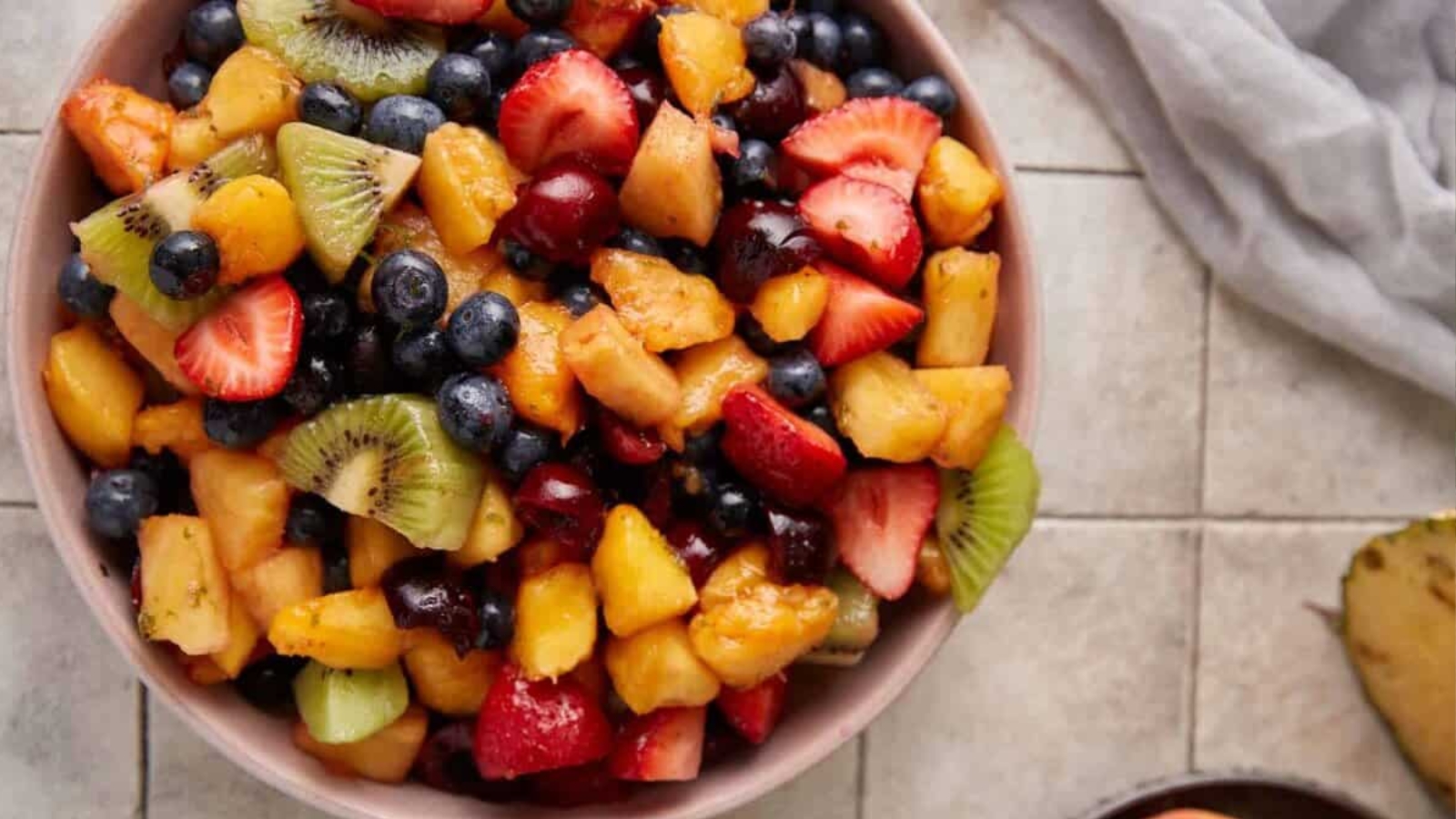A bowl filled with a mixed fruit salad containing strawberries, blueberries, kiwi, melon, grapes, and pineapple pieces, placed on a tiled surface.