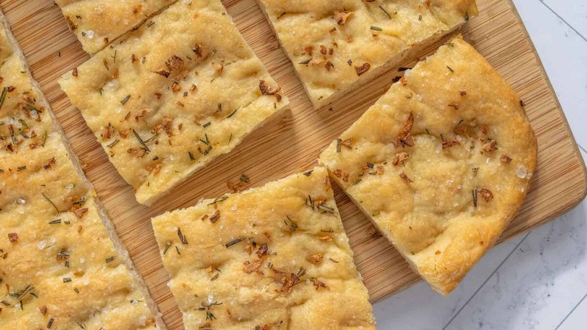 Sliced pieces of golden brown focaccia bread topped with chopped herbs and garlic on a wooden cutting board.