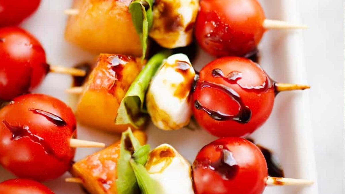 Skewers with cherry tomatoes, mozzarella balls, basil leaves, and cantaloupe pieces, drizzled with balsamic glaze, arranged on a white plate.