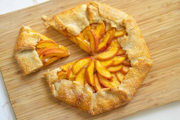 A peach galette with a golden, flaky crust sits on a wooden cutting board, with one slice cut and slightly separated from the rest.