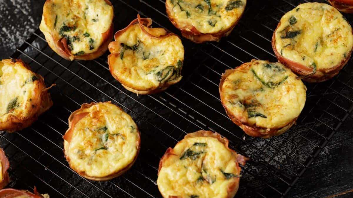 Mini egg frittatas with visible spinach baked in muffin tins, cooling on a black wire rack.