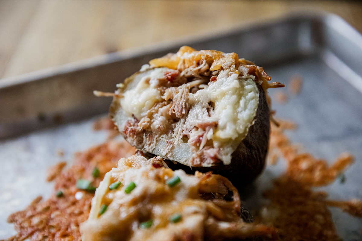 A baked potato cut open, filled with shredded meat and melted cheese, garnished with chopped chives, on a metal tray.