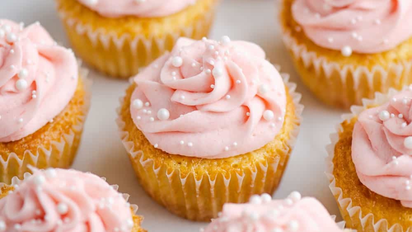 Vanilla cupcakes topped with pink buttercream frosting and white pearl sprinkles, arranged closely together on a white surface.