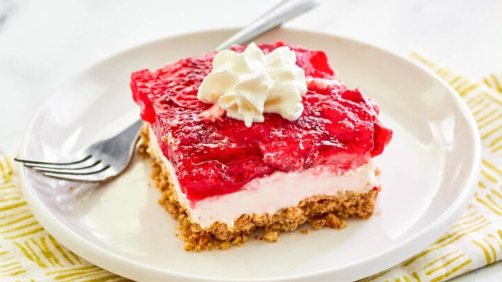 A dessert with a crumbly base, creamy middle layer, and bright red gelatin topping, garnished with a dollop of whipped cream on a white plate next to a fork.