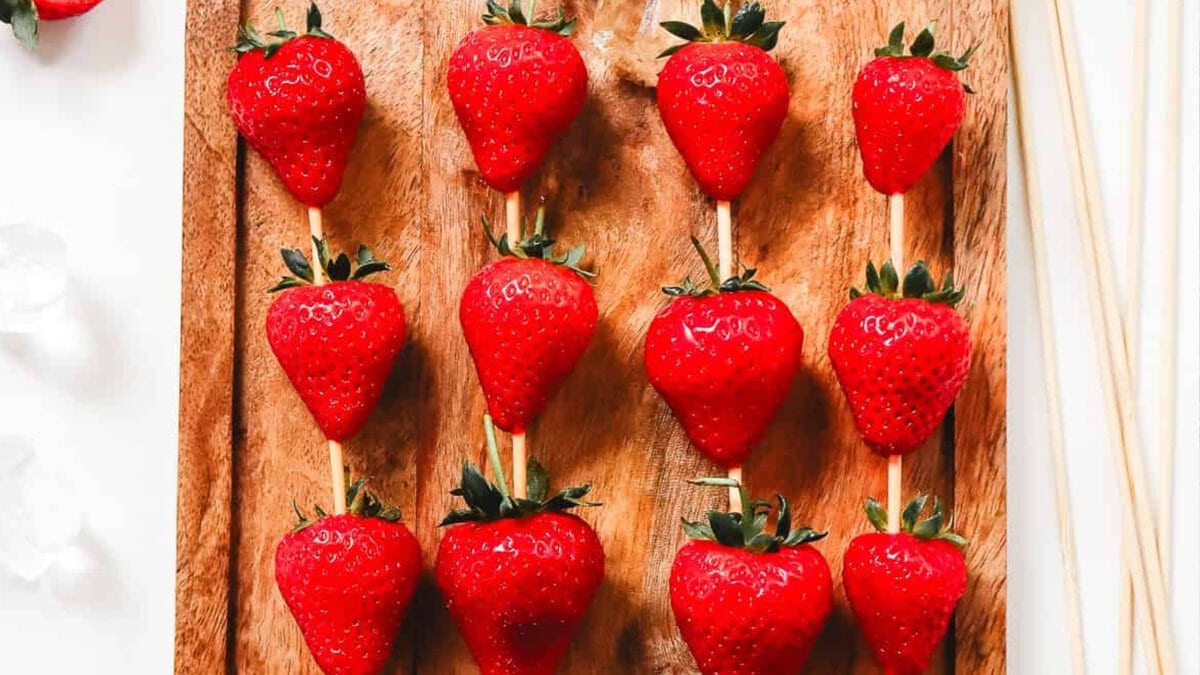 Fresh strawberries and ice cubes on skewers rest on a wet wooden cutting board, with a few ice cubes scattered around.