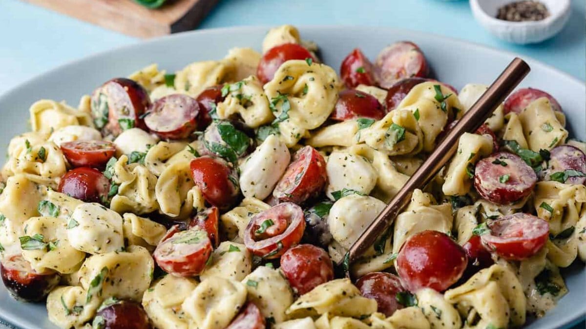 A close-up of a pasta salad with cheese tortellini, cherry tomatoes, mozzarella balls, fresh herbs, and a fork on a blue plate.