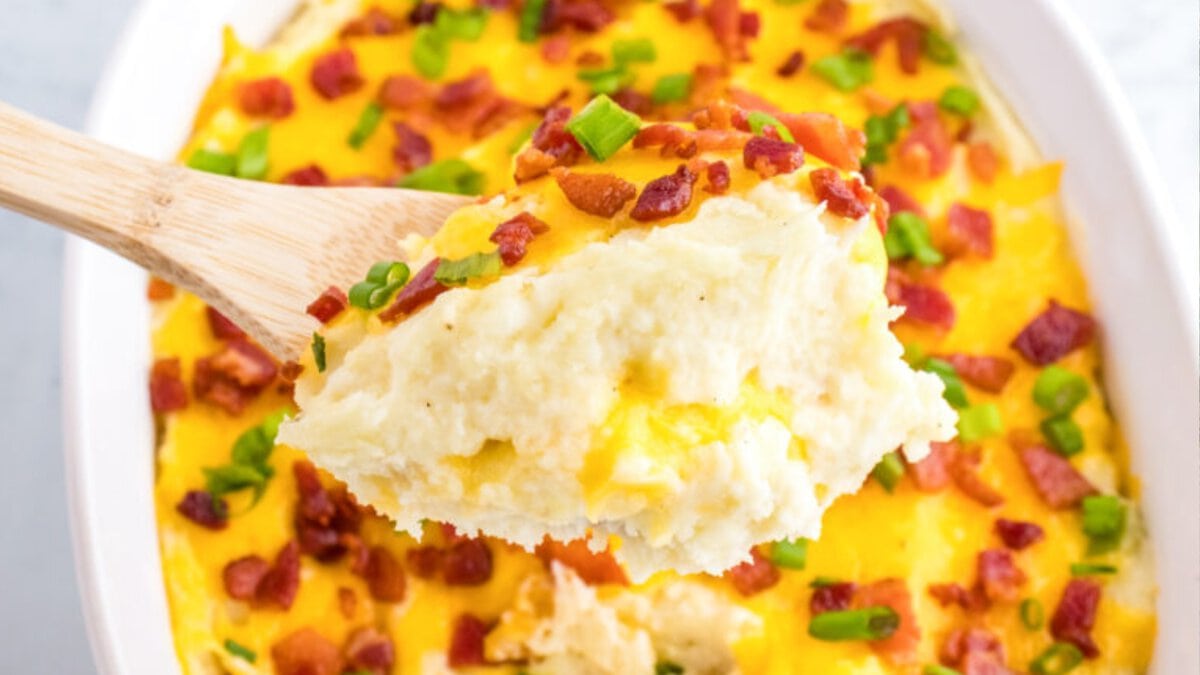 A wooden spoon lifts a serving of cheesy mashed potato casserole topped with bacon bits and chopped green onions from a white dish.