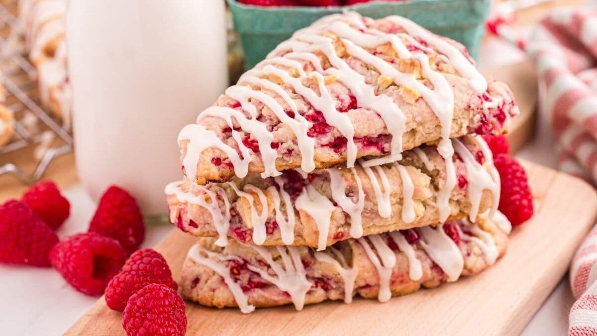 Stack of three White Chocolate Raspberry Scones drizzled with icing on a wooden board, surrounded by fresh raspberries and a bottle of milk.