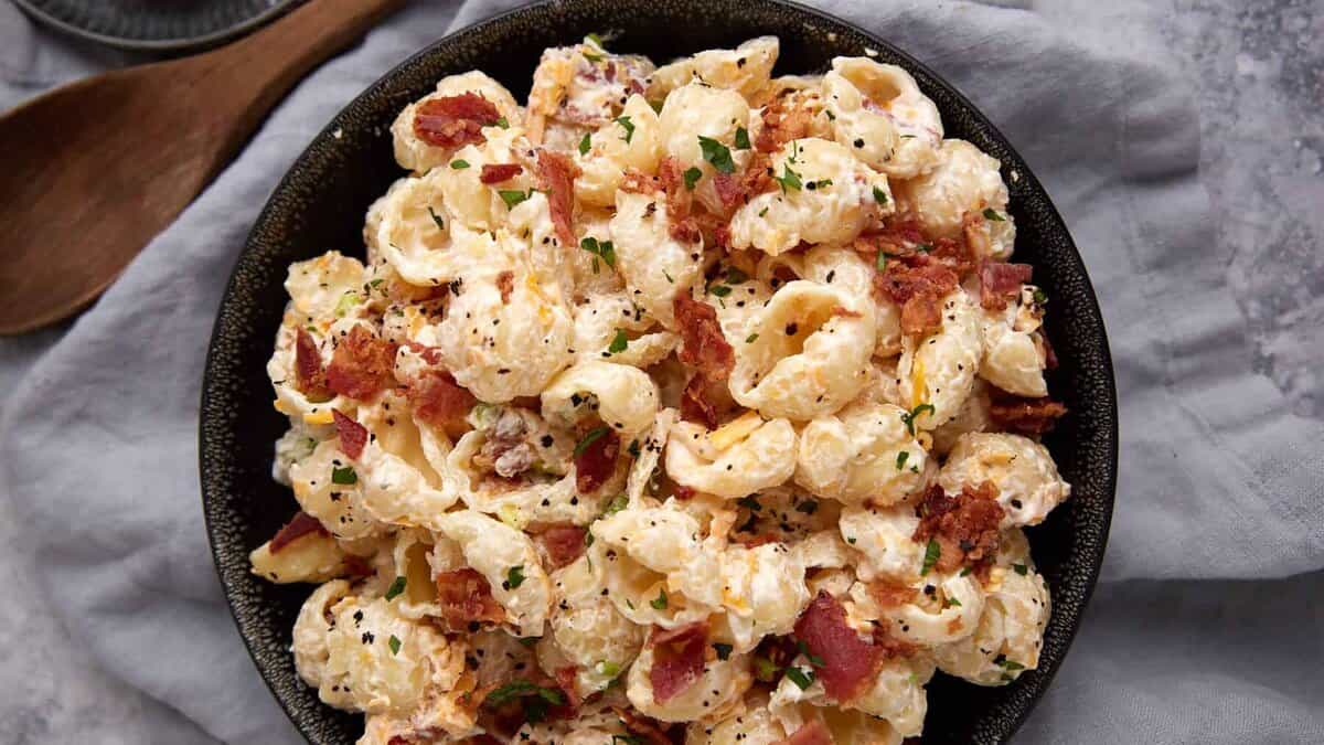 A bowl of creamy pasta shells mixed with bacon pieces and garnished with chopped herbs, placed on a gray cloth.