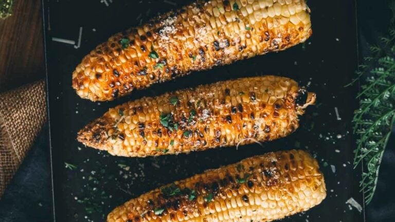 Three grilled corn on the cob pieces are placed side by side on a black surface, sprinkled with herbs and grated cheese.